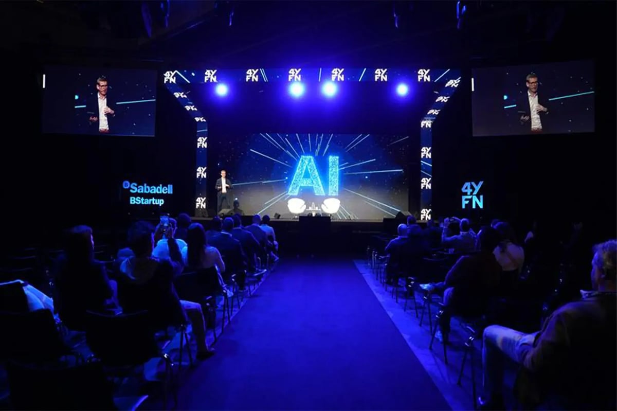 The image depicts a speaker presenting on stage at an event, with a large "AI" graphic displayed in the background. The stage is illuminated with blue lighting, creating a high-tech, futuristic atmosphere. The audience is seated in rows, facing the speaker, and the 4YFN logo is visible on the sides of the stage. The event seems to focus on AI, and the audience appears to be engaged in the presentation. There is also a camera capturing the event from the front. The sponsor "Sabadell BStartup" is displayed on the left side of the stage.