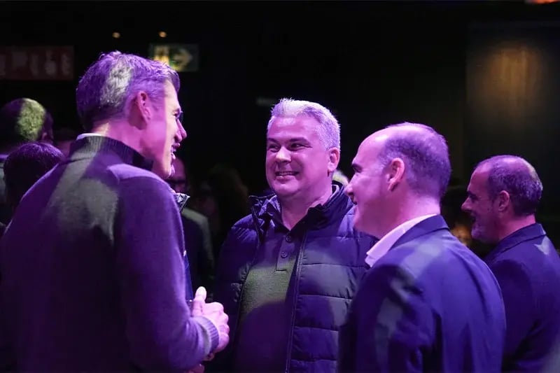 Group of professionally dressed men engaged in friendly conversation under purple lighting at a networking event, smiling and enjoying the informal atmosphere.