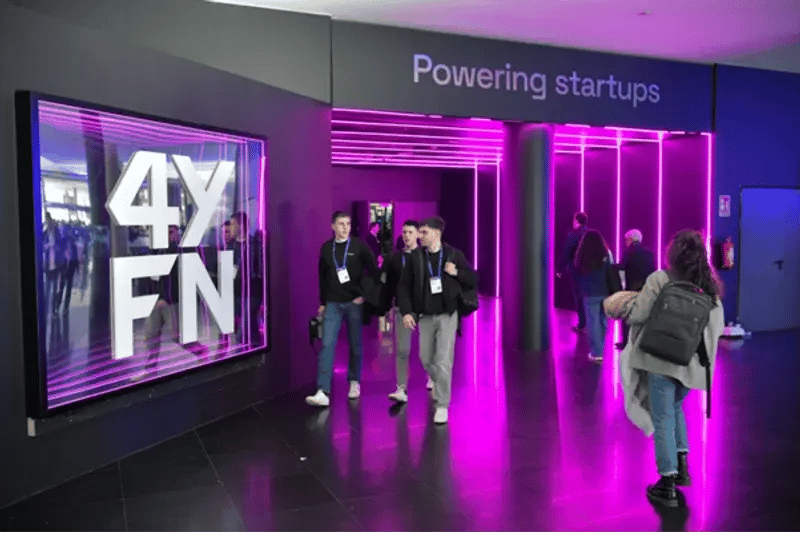 Group of people walk past a black wall with pink neon lights and a 4YFN logo. The sign "Powering startups" conveys an energetic, modern atmosphere.