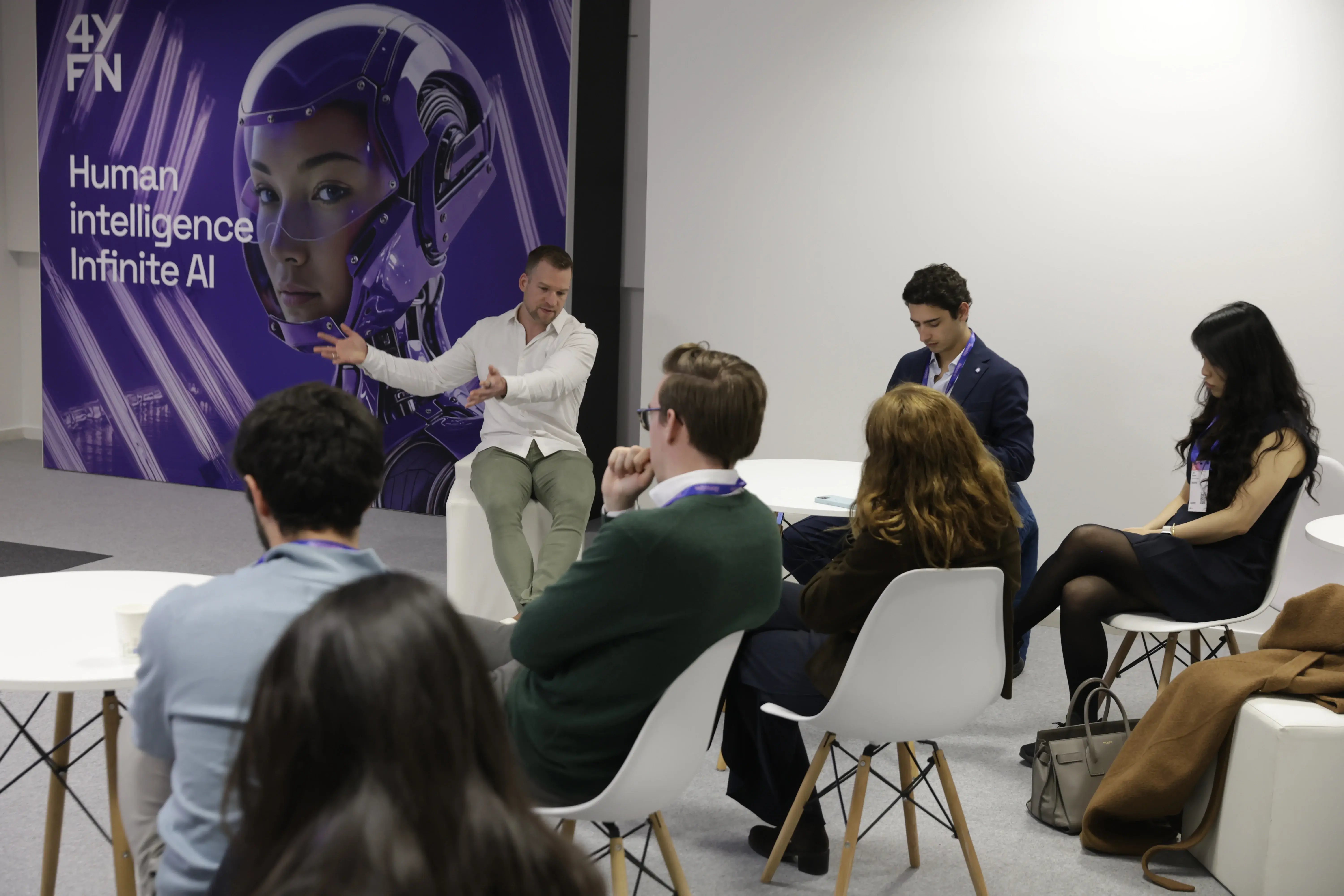 A group of people sits in a seminar setting, attentively listening to a speaker gesturing while speaking. A large futuristic AI-themed poster is visible in the background. The mood is focused and engaged.