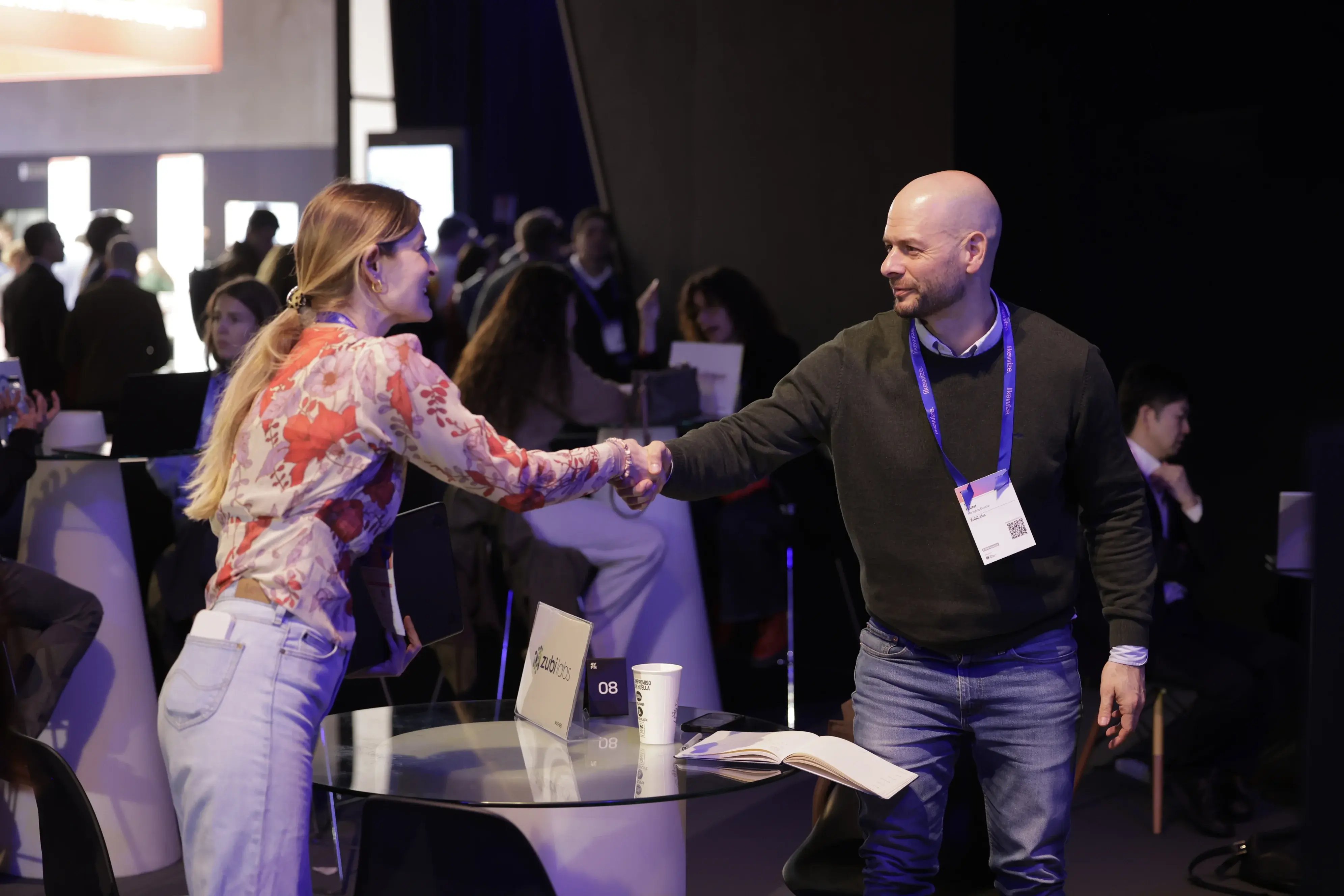 A man and a woman shake hands at a conference table, engaging in friendly conversation. The room is filled with people networking in a professional setting.