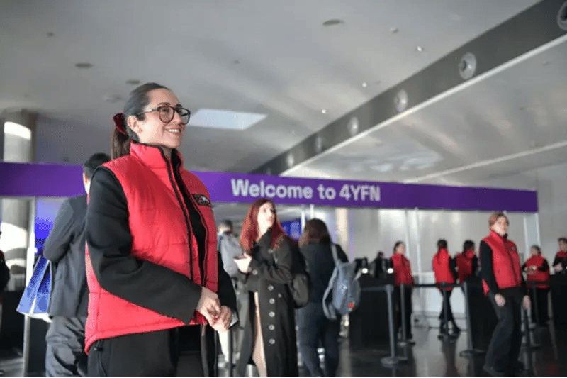 Smiling woman in red uniform standing in front of a "Welcome to 4YFN" sign. People are entering the event, creating a welcoming atmosphere.