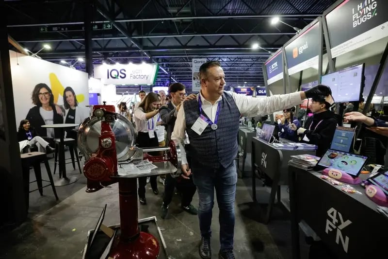 A lively tech expo scene with people interacting at various booths. A man in a vest gestures towards a screen, creating a bustling, engaging atmosphere.