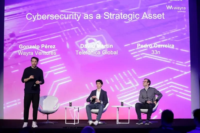 Three men on stage discussing "Cybersecurity as a Strategic Asset." A digital blue and purple background with electronic circuit patterns enhances the tech atmosphere.