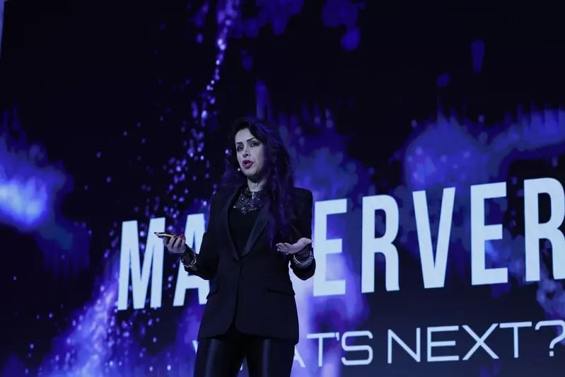 A speaker presenting on stage with a bold purple and black backdrop featuring the text 'Metaverse: What's Next?' in a futuristic style.