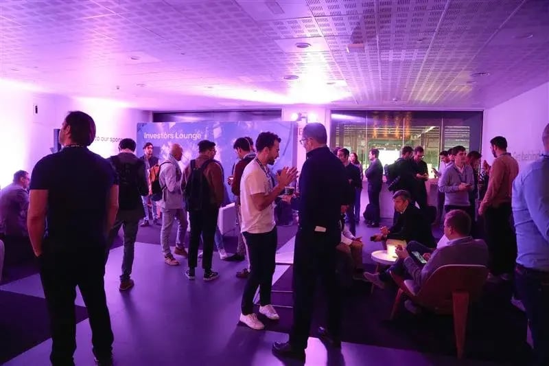 The image depicts the "Investors' Lounge" at the 4YFN (4 Years From Now) event, which is a part of MWC (Mobile World Congress). It shows a group of people gathered and networking in a brightly lit, modern lounge area with purple lighting. Individuals are seen standing and chatting in small groups, while others are seated in lounge chairs, some of them engaged with their devices. A wall in the background has the "Investors' Lounge" sign and displays the event's sponsor. The space appears to be designed for informal networking and discussions among investors and startup founders.