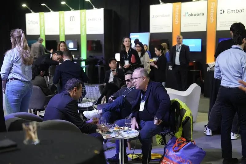The image showcases an exhibition area at the 4YFN (4 Years From Now) event, where various startups are presenting their solutions. Several individuals are engaged in networking and conversations. Some are seated at a table, chatting and sharing food and drinks, while others are standing and interacting with the displays in the background. The booths behind them feature branding from various startups, with signs displaying their names such as "Fintech," "Okora," "Embat," and others. The space appears busy with a mix of casual networking and discussions about business opportunities and innovations.