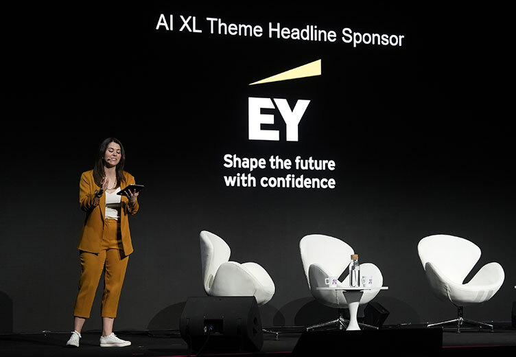 A woman in a mustard yellow suit presents on stage beside a screen displaying the EY logo and the slogan “Shape the future with confidence” during an AI-themed event sponsored by EY. Three white modern chairs and a table with a water bottle are also visible on stage.
