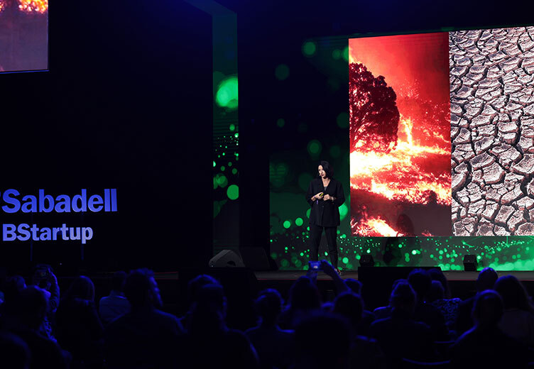 A speaker presents on stage at a Sabadell BStartup event, with a large screen behind them showing images of a wildfire and cracked dry earth, highlighting environmental issues. The audience is seated in the foreground.
