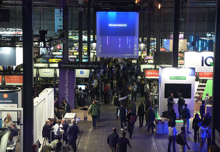 A bustling exhibition hall at a startup event featuring numerous branded booths, including areas labeled "University & Spin-off Hub" and "Pitch Point." Attendees walk through the aisles and engage in discussions under a large hanging screen displaying "Startups stand out!"