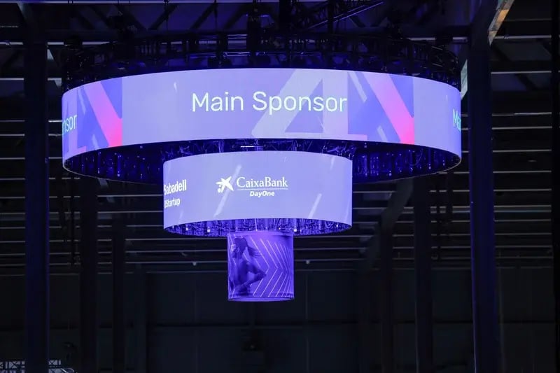 Hanging circular display with "Main Sponsor" text, lit in blue and purple hues. Logos of CaixaBank and Sabadell visible. Tech event ambiance.