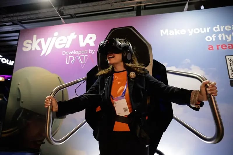 A woman wearing a VR headset smiles, holding onto handles of a VR flight simulator at a tech event. The backdrop displays "skyjet VR."